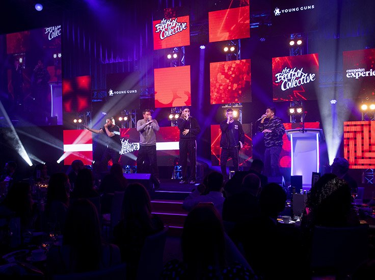 A beatboxing group perform on a big stage with lots of lights, red led panels behind them and an audience in silhouette sitting in front